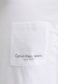 Vit bomull t-shirt med en liten bröstficka. Texten på fickan lyder "Calvin Klein Jeans sedan 1978" i svart. Slät textur.