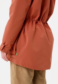 Giacca impermeabile rossa con vita regolabile e dettagli elastici. Presenta una texture liscia e un logo Vaude discreto vicino all'orlo.