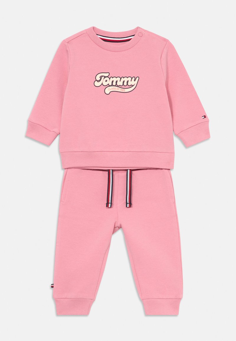 Sweater rosa com o logótipo "Tommy", gola redonda e botões de pressão. Calças de fato de treino rosa em conjunto com cordão às riscas e punhos canelados.