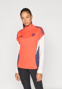 Élénk narancssárga sportpulóver magas gallérral, fehér hosszú ujjakkal, tengerészkék oldalpanelekkel és hímzett logóval a mellkason.