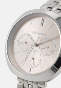 Calvin Klein sølvfarget klokke med en rund, lys rosa urskive som har tre sub-dialer, minimalistiske timeindekser og en armbånd i rustfritt stål.