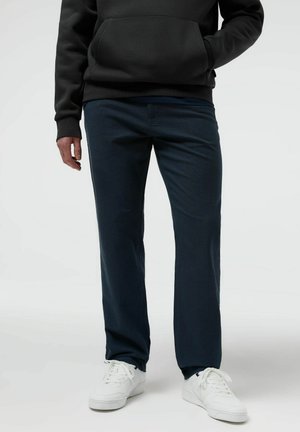 Osoba ubrana w ciemnoniebieskie spodnie o prostych nogawkach, czarną bluzę z kapturem oraz białe trampki, stojąca z jedną ręką w kieszeni, a drugą swobodnie opuszczoną.