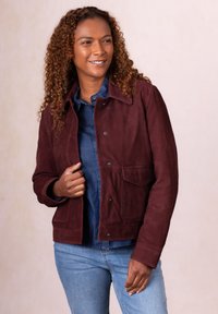 Woman with curly hair wearing a burgundy buttoned jacket over a blue shirt and light blue jeans, smiling and looking to the side.