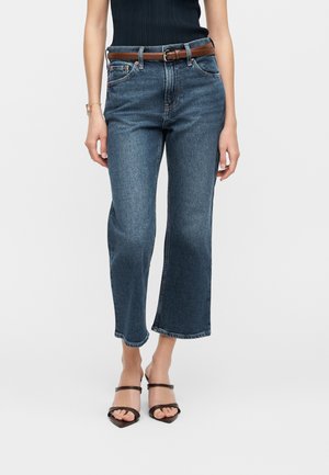 Žena má na sebe kratšie modré rovné rifle, hnedý opasok, čierne tielko bez rukávov a čierne sandále na opätku so remienkami.