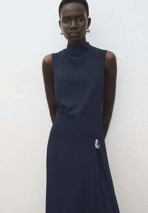 Vestido sin mangas de color navy con cuello alto, que presenta un diseño drapeado y un dobladillo asimétrico. Los detalles incluyen un brillante acento plateado cerca de la cintura.