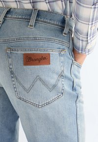 Vaqueros de mezclilla azul claro con un bolsillo trasero que lleva un parche con el logo marrón de Wrangler, combinados con una camisa de cuadros azul y blanca.