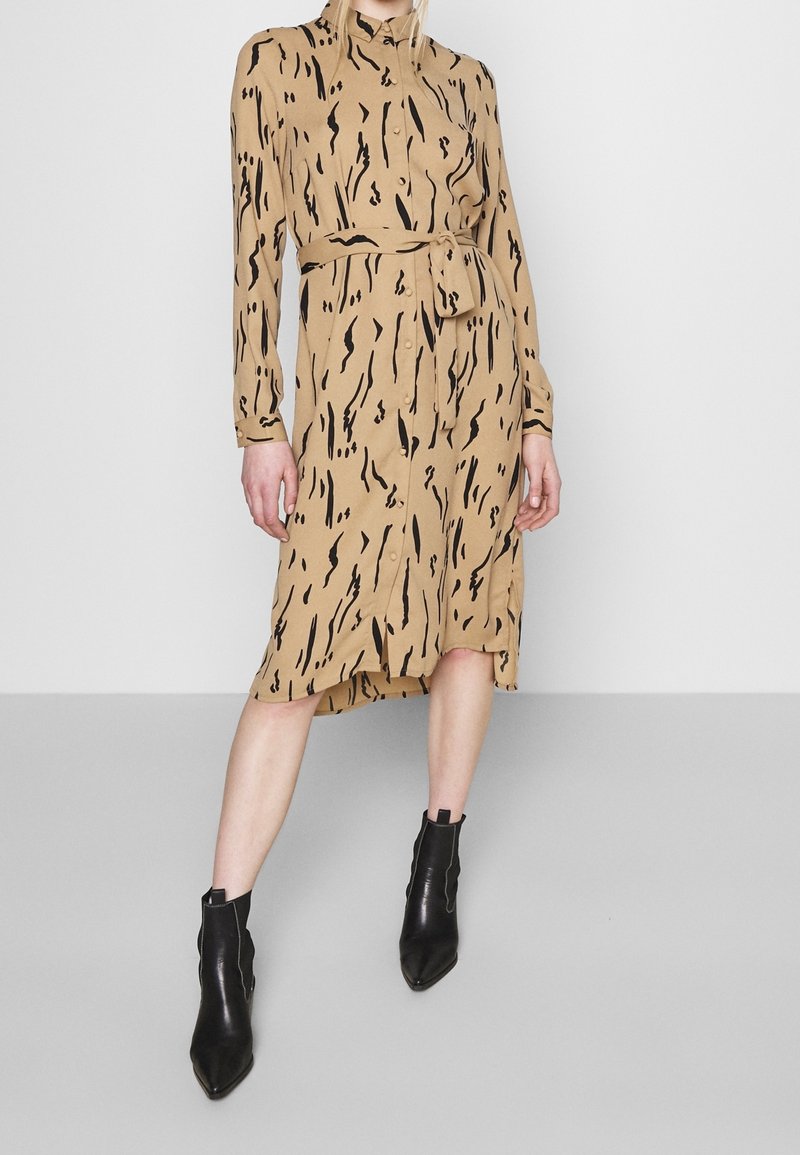 Robe chemise midi beige avec imprimé abstrait noir, manches longues, boutonnée devant, ceinture à la taille, portée avec des bottines noires.