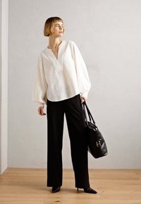 Blusa bianca con motivo sottile e scollatura a V, abbinata a pantaloni neri a gamba larga. La modella tiene una borsa nera a spalla, in piedi su uno sfondo chiaro.