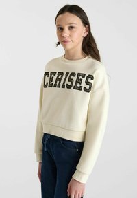 Crèmekleurig cropped sweatshirt met vetgedrukte zwarte tekst "CERISES". De stof lijkt zacht en heeft ribboordjes aan de mouwen en onderkant, gecombineerd met donkerblauwe jeans.
