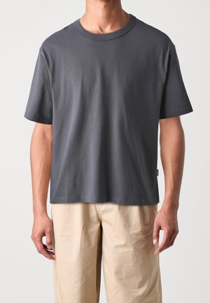 Person iført en ensfarvet mørkegrå kortærmet T-shirt og lyse beige bukser, stående mod en ensartet lys baggrund.