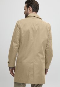 Trench-coat beige avec un col classique, des manches longues et des poignets boutonnés. Il comprend une fente à l'arrière et un discret logo de la marque au niveau du col.