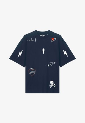Jeune personne portant un t-shirt bleu marine avec des symboles graphiques blancs et rouges, un pantalon cargo noir et des baskets noires, debout sur fond blanc.
