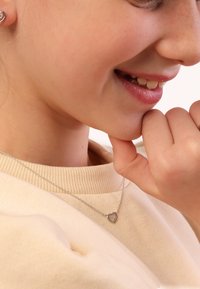 Zilveren hartketting met een gestructureerd ontwerp, voorzien van kleine heldere stenen. Draagt op een licht truitje, waardoor de delicate ketting goed tot zijn recht komt.