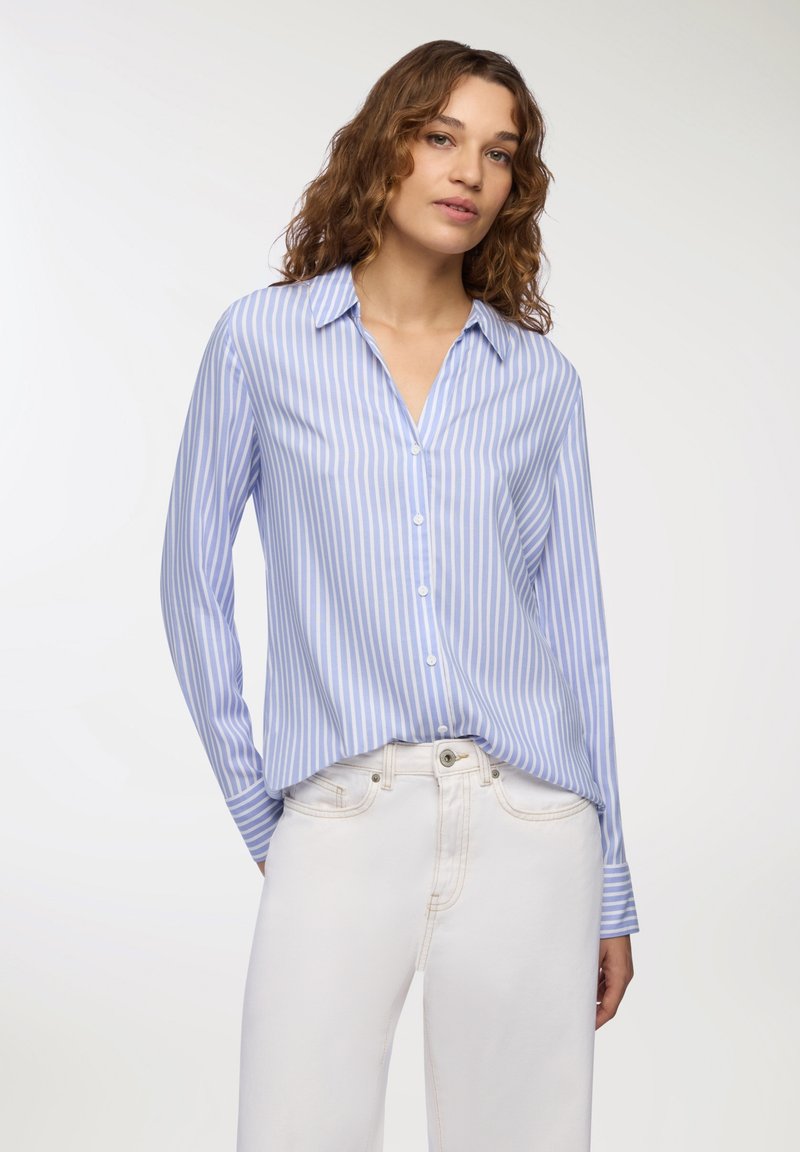 Camicia blu chiaro con bottoni, righe bianche verticali, maniche lunghe e colletto a punta, abbinata a pantaloni a gamba larga color crema.
