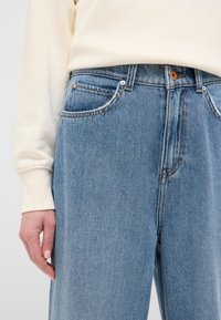 Jeans in denim azzurro chiaro con un taglio rilassato, cuciture visibili e chiusura con bottone. Sono evidenti tasche anteriori e una texture liscia.