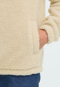 Lys beige fleece pullover med en tekstureret, fluffy overflade. Har ærmer med manchetter og en sidelomme, parret med mørkeblå jeans.