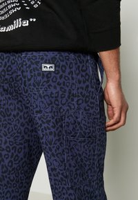 Muž v tmavě modrých kalhotách s černým leopardím vzorem a několika kapsami, doplněný o černou mikinu s částí bílého textu.