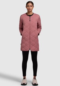 Veste longue matelassée rose avec un col rond, fermeture éclair et poches latérales. Portée sur des leggings noirs et des chaussures de sport de couleur claire.