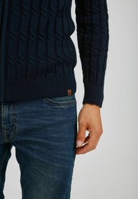 Marineblå strikket cardigan med et kabelmønster, ribbet manchetter og kant. Båret over denim jeans, med et lille læder mærke nær bunden.