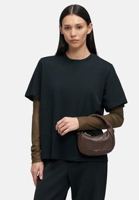 Kobieta o długich ciemnych włosach nosi czarny, oversize'owy t-shirt na brązowych długich rękawach, trzyma małą brązową torebkę z krótkim uchwytem.