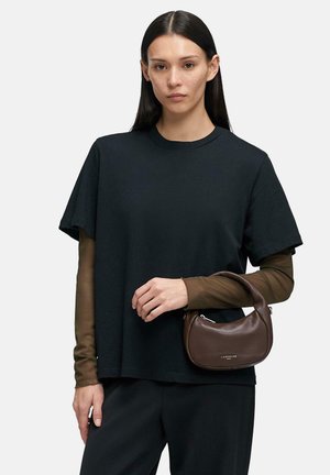 Kobieta o długich ciemnych włosach nosi czarny, oversize'owy t-shirt na brązowych długich rękawach, trzyma małą brązową torebkę z krótkim uchwytem.