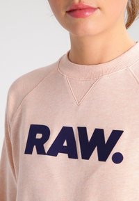 Světle růžová mikina s kulatým výstřihem, na přední straně s velkým námořnickým modrým nápisem "RAW." Vyšívaná tkanina s žebrovanými detaily.