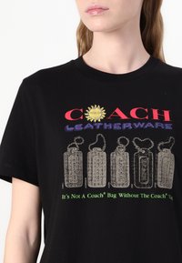 Camiseta de algodón negra con el logotipo "COACH LEATHERWARE" en rojo y púrpura. Gráfica de etiquetas colgantes en gris con texto en verde debajo.