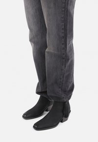 Botines negros con diseño de punta afilada, que cuentan con un pequeño tacón cuadrado y un borlas decorativas en la cremallera. Llevados con jeans grises.