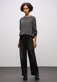 Donna con capelli corti e ricci, che indossa una camicia a maniche lunghe a righe bianche e nere, jeans neri a gamba larga e stivali neri.
