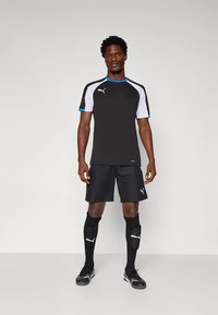 Camisola desportiva preta com detalhes em branco e azul, combinada com calções pretos e meias até ao joelho. Apresenta o logótipo da Puma na roupa e no calçado.