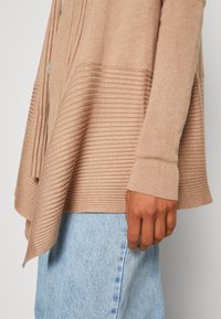 Haut à manches longues marron clair côtelé avec ourlet asymétrique, porté avec un jean en denim bleu clair, montrant le côté et la main d'une personne.
