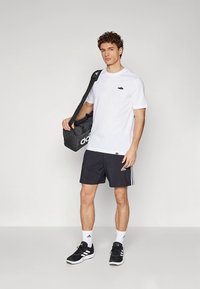 Hvit t-skjorte med svart logo, svarte shorts med hvite sidestriper, svarte sportssko og en svart duffelbag. Enkel, atletisk design.