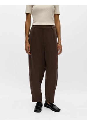 Person trägt lockere dunkelbraune Hose mit umgeschlagenen Säumen, beige kurzärmliges Oberteil und schwarze glänzende Loafers, steht vor einem schlichten Hintergrund.
