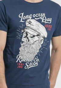T-shirt en coton bleu marine arborant un graphique blanc d'un crâne avec une barbe et un chapeau de capitaine, avec le texte "Long Ocean Day" et "Ritchie Co." en rouge.