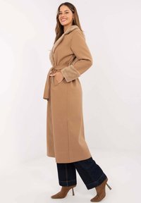 Langer, kamelfarbener Mantel mit Pelzkragen und -manschetten, tailliertem Gürtel, Seitentaschen und gerader Silhouette. Getragen über weiten, dunklen Jeans.