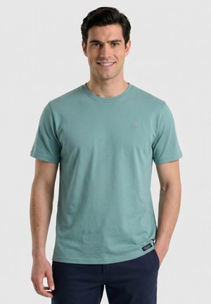 Hombre sonriente que lleva una camiseta de cuello redondo color verde azulado y pantalones oscuros, de pie con una mano en el bolsillo frente a un fondo liso y claro.