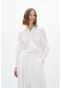 Weiße Bluse mit V-Ausschnitt, versehen mit bestickten Details und plissierten Ärmeln, kombiniert mit hellen, hochtaillierten Hosen.