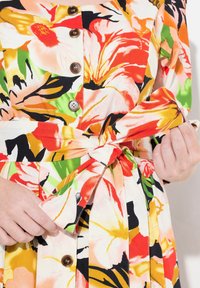 Blumenkleid mit einem lebhaften Muster in Orange, Pink und Grün. Verfügt über eine taillierte Taille, Knöpfe und Faltenrockdetails. Hergestellt aus leichtem Stoff.