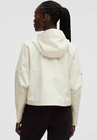 Kort vit regnjacka med huva, med en slät yta, breda ärmar och axelsömmar. Baksidan visar minimala design detaljer.