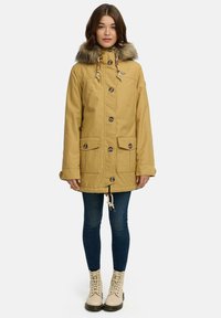 Żółta kurtka parka z futrzanym kapturem, zapięcie na guziki, sznurek ściągający i dwie przednie kieszenie. Noszona na dżinsy i beżowe buty na sznurówki.