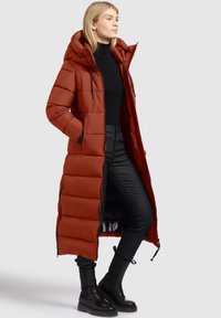 Vrouw in een lange roestkleurige pufferjas met capuchon, zwarte turtleneck, zwarte leren broek en zwarte enkellaarsjes, staand met haar handen in haar zakken.