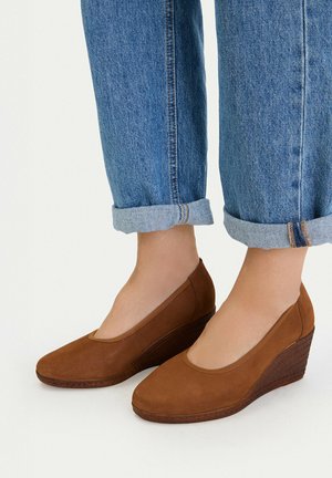 Pies con zapatos cuña marrones combinados con vaqueros azules con puños contra un fondo liso.