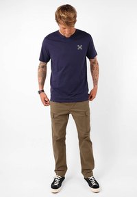 T-shirt en coton bleu marine avec un petit motif sur la poitrine, associé à un pantalon cargo beige et des baskets noires avec des semelles blanches.