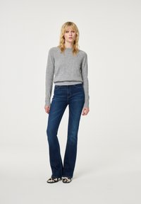 Grijze gebreide trui met lange mouwen, gecombineerd met donkerblauwe flared jeans. Model draagt zwart-witte gepatterneerde schoenen en staat op een neutrale achtergrond.