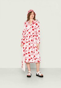Vestido longo rosa com padrões de flores vermelhas, colarinho, frente com botões, mangas longas; combinado com um chapéu de balde vermelho e branco a condizer e sandálias pretas.