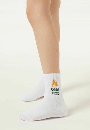 Gambe con calzini bianchi decorati con una grafica di fiamme e la scritta "COOL KID" sul lato, in piedi su un pavimento chiaro.