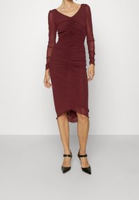 Figurované bordo šaty s dlouhými průsvitnými rukávy, nabíranou texturou a lemováním, které mírně klesá vzadu, spárované s černými špičatými podpatky.