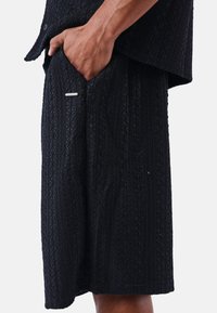 Pantalones cortos negros con un tejido texturizado y estampado, que cuentan con un bolsillo lateral y un pequeño acento metálico cerca de la abertura del bolsillo.