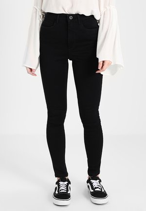 Personne portant un jean noir slim, un chemisier blanc à manches longues évasées et des baskets noires et blanches à lacets.