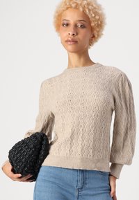 Beige knitted sweater with a wavy pattern and ribbed cuffs, paired with a textured black handbag. The sweater features a crew neck design.
